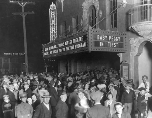 A black and white photo of a large crowd from 1923 standing outside a building which has a sign which says Bard and a large marquee which says Baby Peggy in "Tips"