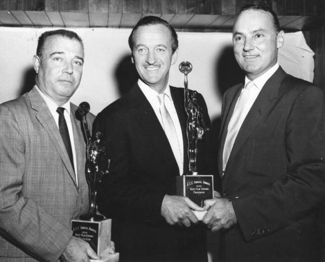 Three men with light-tone skin are dressed in suits. Two awards statuettes are being held.