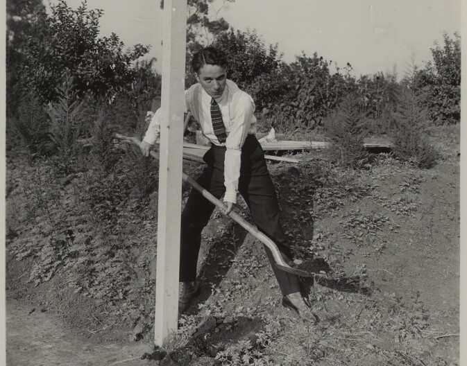 A black and white photo of a white man in a tie shoveling dirt.