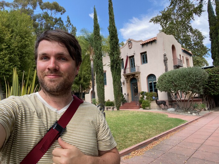 A smiling man with brown hair, a beard, a striped t-shirt and a red bag strap across his chest takes a selfie of himself with a home in the background. The home is a two-story Spanish colonial style house with arched windows, a tiled roof, a lawn and landscaped trees out front.