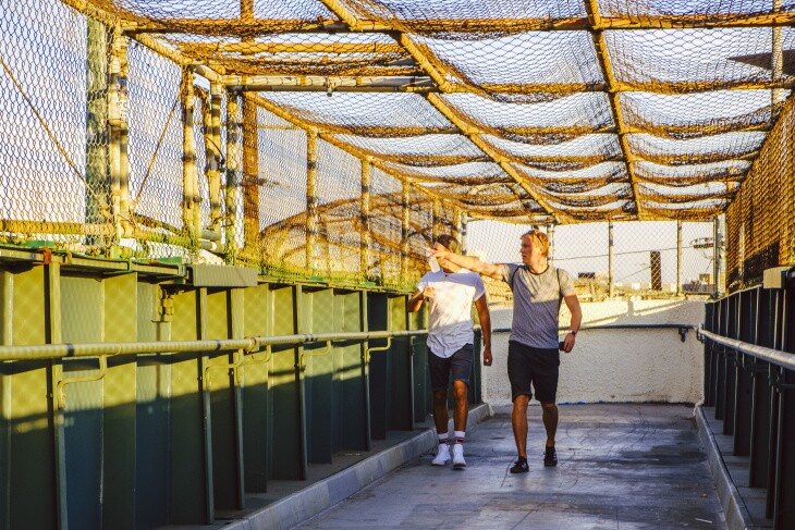 Two pedestrians look off into the distance -- presumably at all the traffic -- as they walk through a fenced in overpass that traverses the 110 freeway, and heads to the ballpark.