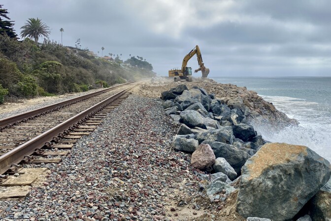 Looking down a train track with waves crashing into boulders on the right hand side, a slope with palm trees and houses on top on the left side, and an excavator about to pick up a boulder next to the tracks in the distance. 