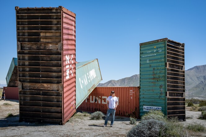 The artist Matt Johnson, wearing a white T-shirt and blue jeans, stands in front of several large metal shipping containers at variously odd angles. Two are green and two are red, and all of them are rusted to different degrees.