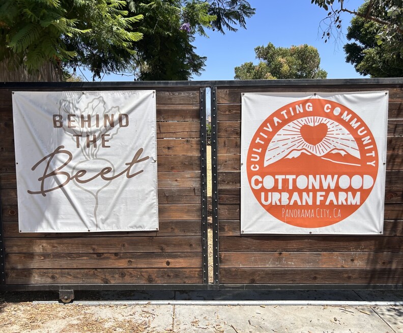 A large wooden gate with black metal trim is latched at the center on a sunny, clear morning. Two signs with logos are affixed to each side. The left is white with brown text that reads "Behind The Beet" and the one on the right is white with orange text that reads "Cultivating Community Cottonwood Urban Farm Panorama City, CA"