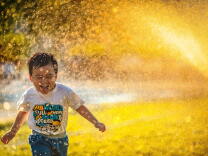 A young Asian child runs through a sprinkler wearing a t-shirt and jeans. He is laughing and the sunshine is bright behind him, illuminating the water. 