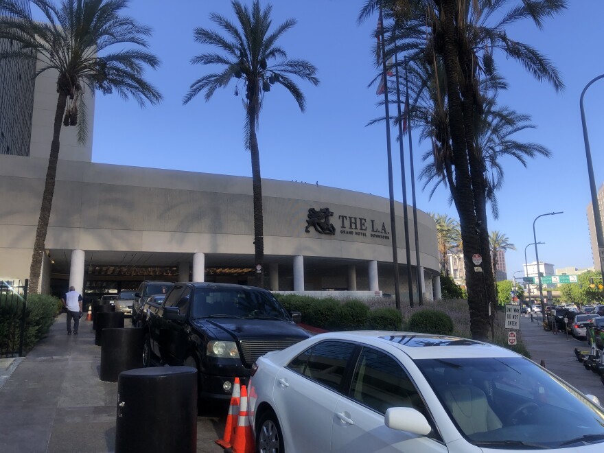 Cars are parked in front of a curved building facade with a sign reading: the L.A. Grand Hotel. Palm trees surround the hotel.