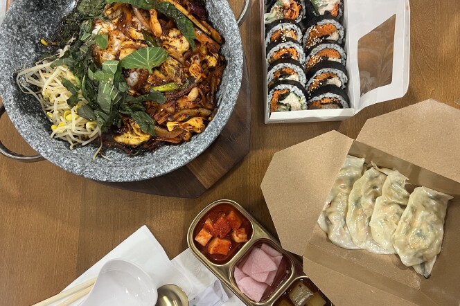 A grey stone pot with caramel colored squid and onions alongside green perilla leaves and yellow bean sprouts. A container open with tightly rolled kimbap and a cardboard box opened to reveal neatly shaped dumplings. There is a container with pickled pear in a fiery red color, pickled radish in soft pink. 