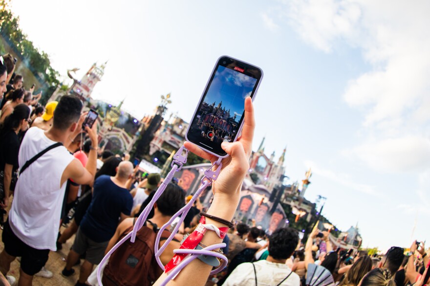 An outdoor music festival space, but the camera is focused on a woman's hand holding up her smartphone to record a video. A light purple lanyard is connected to the phone case and wrapped around her right wrist.
