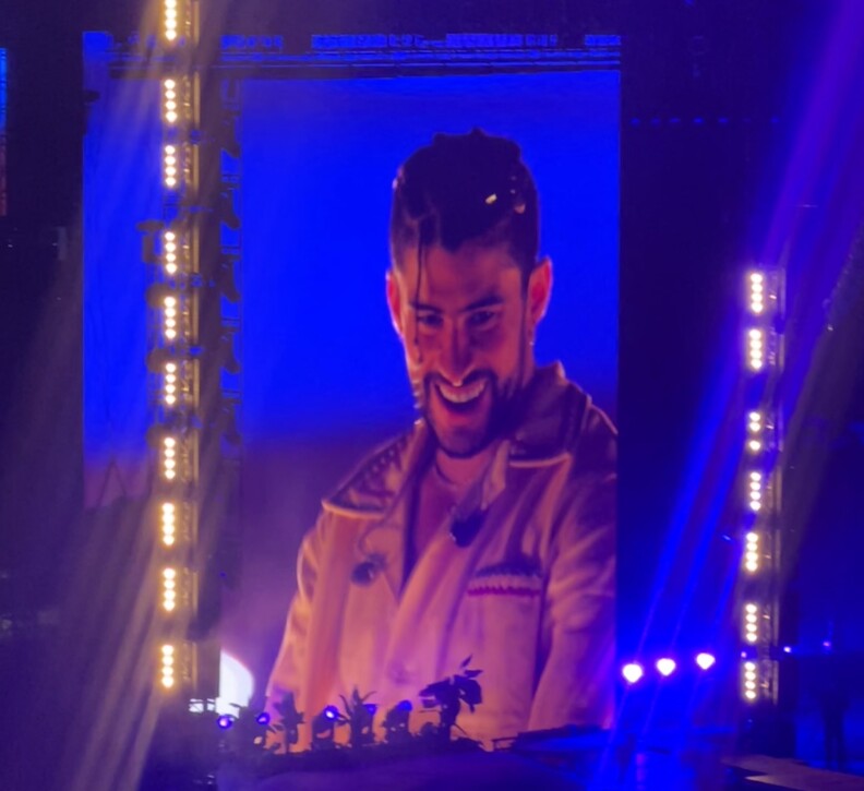 Bad Bunny, wearing a braid in front of his face, smiles at the camera during his concert at SoFi Stadium. The venue is lit in blue with spots of yellow lights that are next to the big screen.
