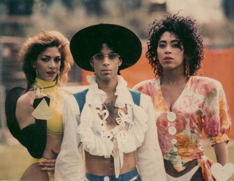 Three people standing outdoors, two women and a man. The man in the center wears a black hat, a white shirt with decorative elements and blue pants. The woman on the left is dressed in a yellow outfit with black accents, and the woman on the right wears a colorful floral top with large buttons.