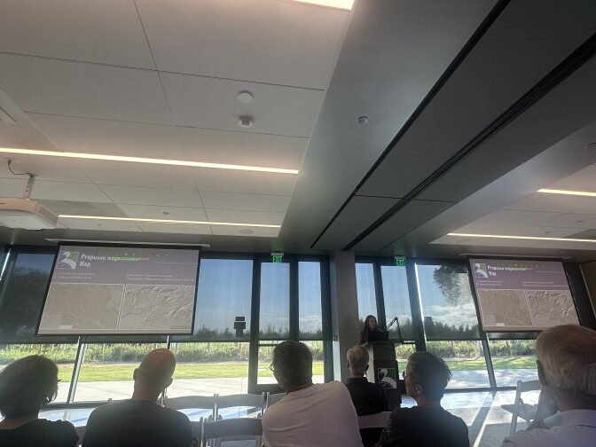 The interior of a community center meeting happening on a sunny day: Many residents are in folding chairs, a person is at a podium and there are two screens at the front of the room. 