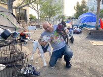 A smiling man wearing a gray t shirt and blue jeans is holding two dogs while kneeling on the sidewalk in the middle of an encampment for unhoused people. 