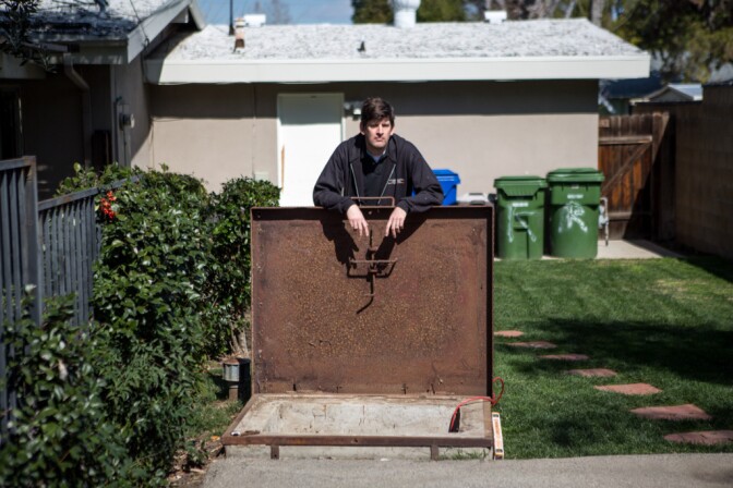 A rusty wide square door opens to a concrete base. A man stands in a yard holding it open. There's green grass and garbage cans behind him.