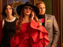 The actress Lindsay Lohan, a white woman who appears to be in her 20s or 30s, is shown in a movie still wearing an expensive-looking red dress, a large black hat. and sunglasses. She is speaking on a cell phone and trailed by a man and a woman. 