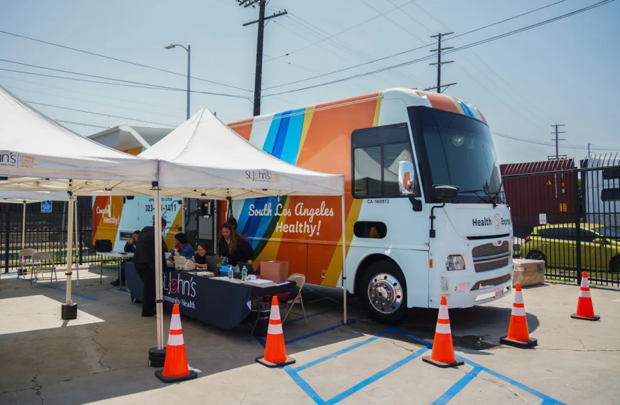 A colorful bus is parked with two white tents standing beside it. People are standing and sitting around a table.