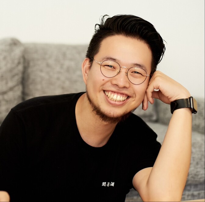 Photo of smiling Taiwanese American man with glasses, resting the side of his face on the back of his hand. 