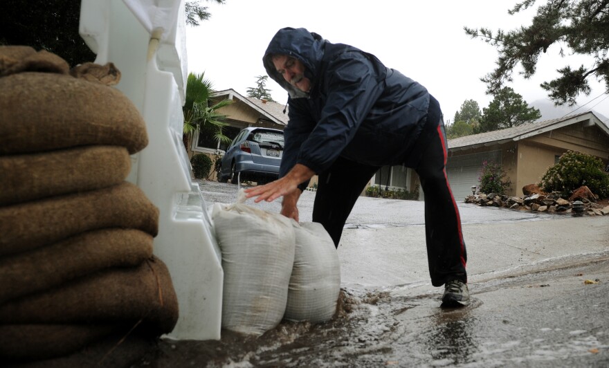 A white man with a gray mustache wearing track pants and a blue rain jacket with hood stands at the driveway of a home placing sandbags against a plastic barricade as water streams and overflows the gutter. Water wells up and flows around his left sneaker.