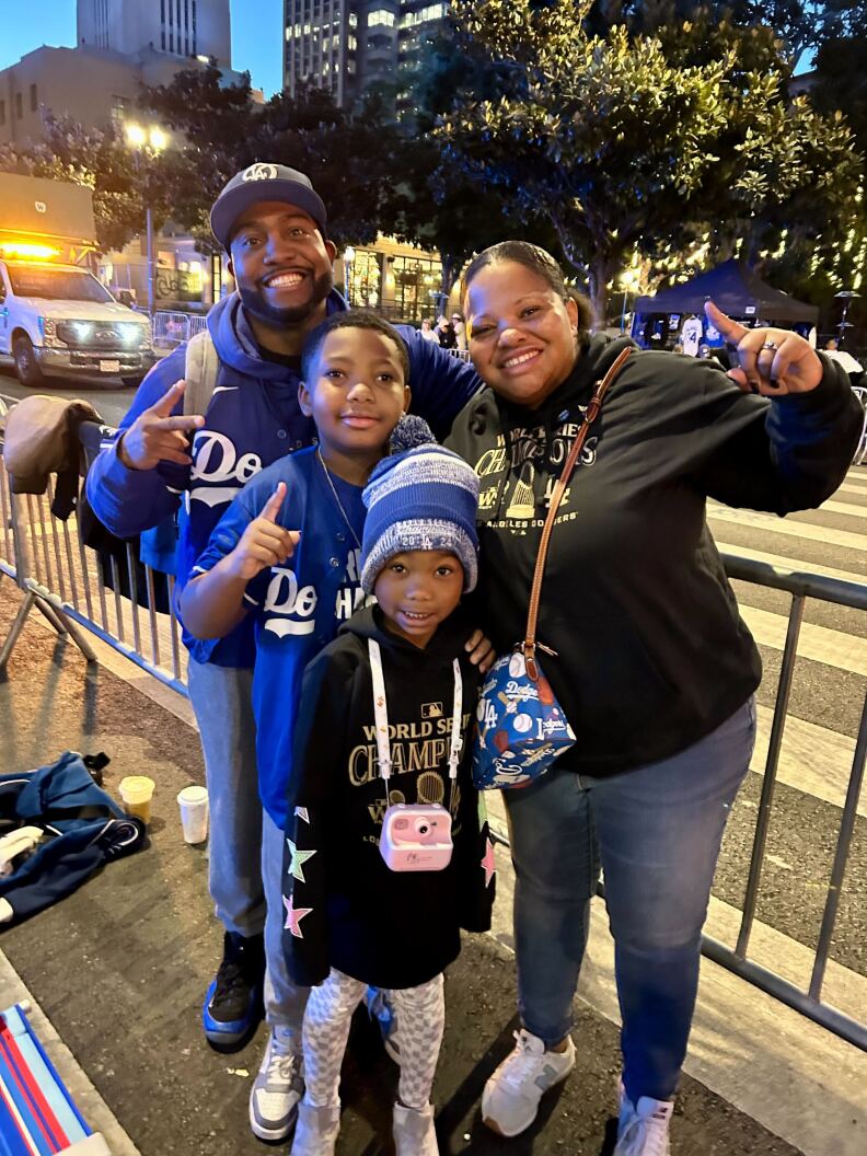 A man with dark skin tone wearing a Dodgers hat and sweatshirt stands next to a woman with medium-dark skin tone wearing a black sweatshirt. In front of them two small children pose wearing Dodgers gear. They stand in a sectioned of area of a street.