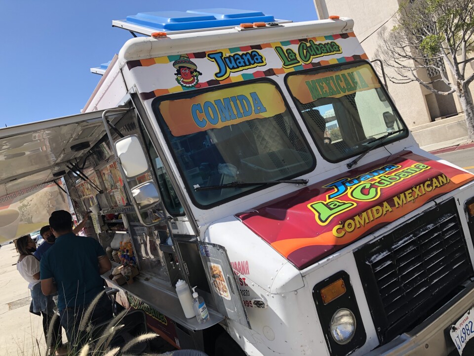 A food truck called "Juana La Cubana" is parked and serving people on a city street on a sunny, clear day.