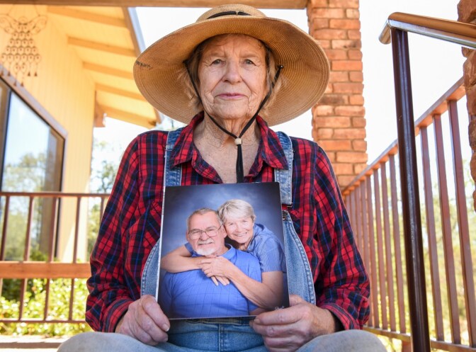 An older white woman holds an approximately 8 by 10 professional photo of herself and her husband smiling together at the camera. She sits outdoors on the steps of a porch and wears denim overalls, a red and blue checkered flannel shirt, and a straw sun hat with a black strap around her chin. 