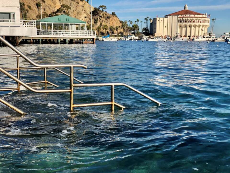 A staircase mostly submerged in ocean water. In the background, a marina and rocky brown hills are visible.