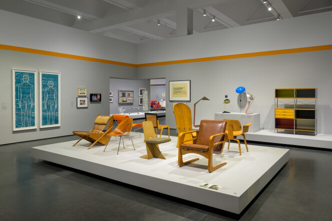 Gallery view of mid-century modern chairs in blond wood and leather displayed on a white platform, with two blue anatomical blueprint-style drawings on the left wall and a colorful modular shelving unit and lamps on a pedestal at right.