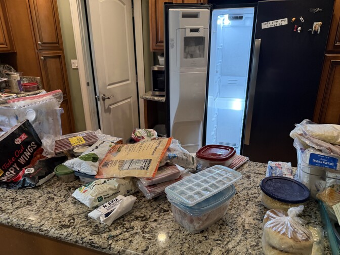 A kitchen counter is cluttered with items like bags of spinach, english muffins, chicken wings and hot dogs. In the background is an empty freezer in a side-by-side fridge.