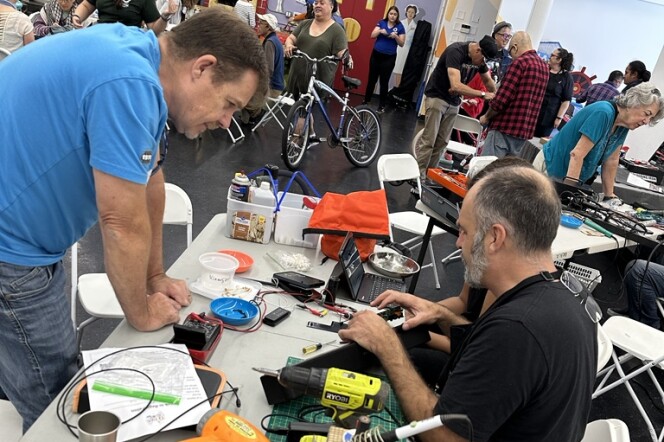 A room full of men and women of all ages. A white foldable table is set up closer to the camera, with two people seated behind it, including a man in blue jeans and a black t-shirt. Another man wearing a blue t-shirt and jeans is leaning over the table, which is covered in tools and electrical equipment.