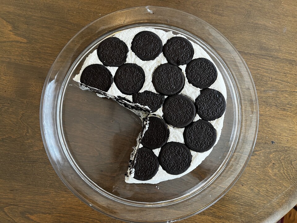 An overhead image of a cake made up of white cream and brown oreos. The cake sits on a clear glass platter on a wood table surface.