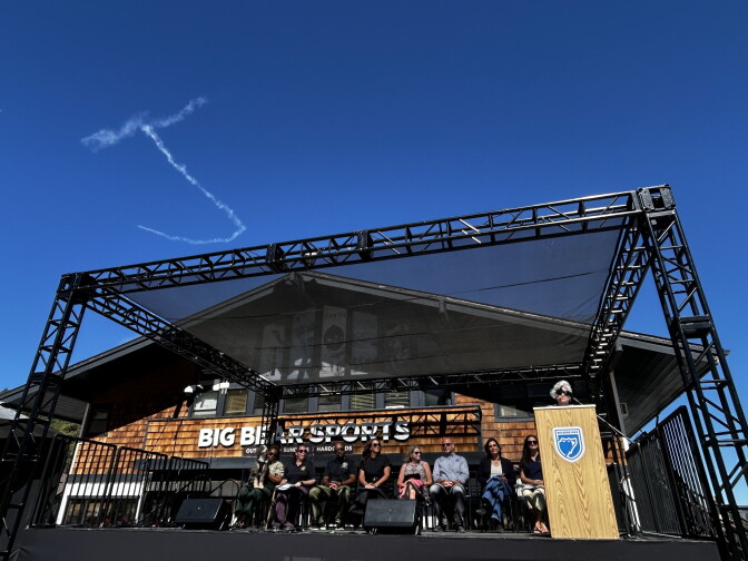 People sit on an outdoor stage in front of the Big Bear Sports building as a person speaks at a podium under a large canopy.