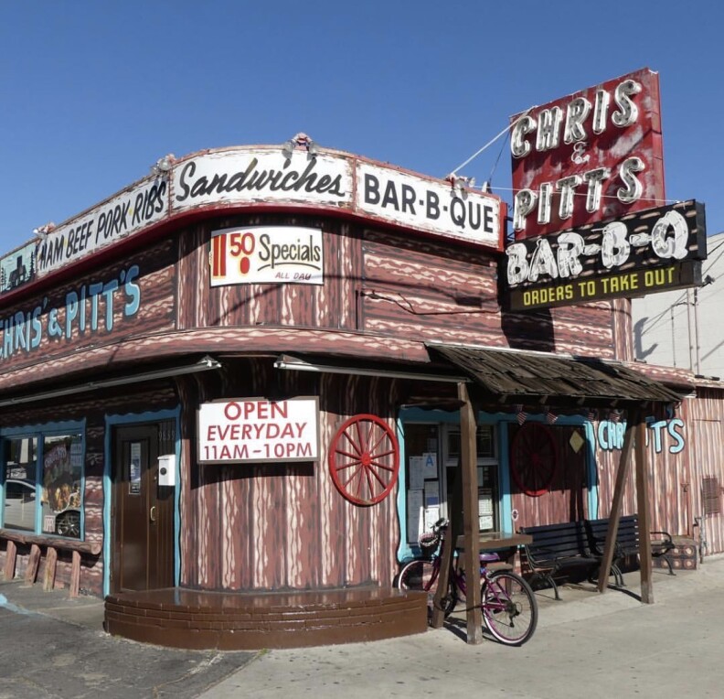 The exterior of a brown building made to look like a log-cabin. The sign reads "Chris' & Pitt's Bar-B-Q. Ham, Beef, Pork, Ribs, Sandwiches."