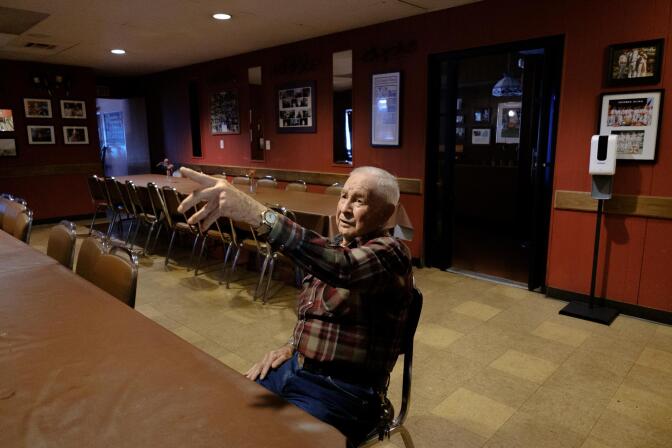 a man in a plaid shirt sits inside an empty dining room and points at something