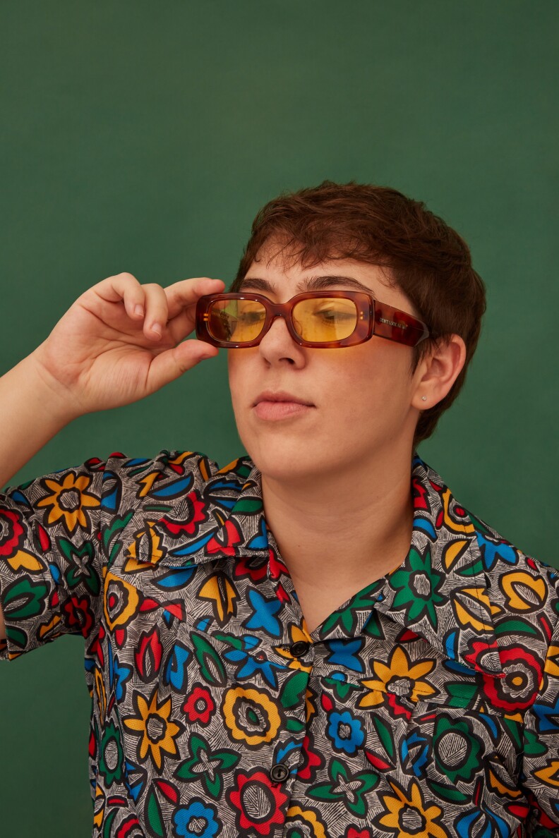 Danielle, a white masculine person with short dark hair and sunglasses, as they hold the frames and look down in front of a solid green background. They're wearing a vibrant shirt with flower shapes.
