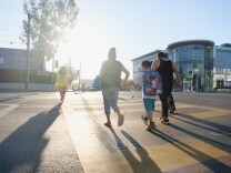 Pedestrians walk in a crosswalk at a busy intersection as a crossing guard walks ahead of them on a bright morning.