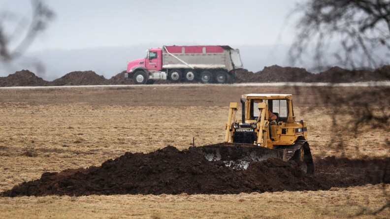 A bulldozer overturns dirt in a field as a truck passes on a road behind it.