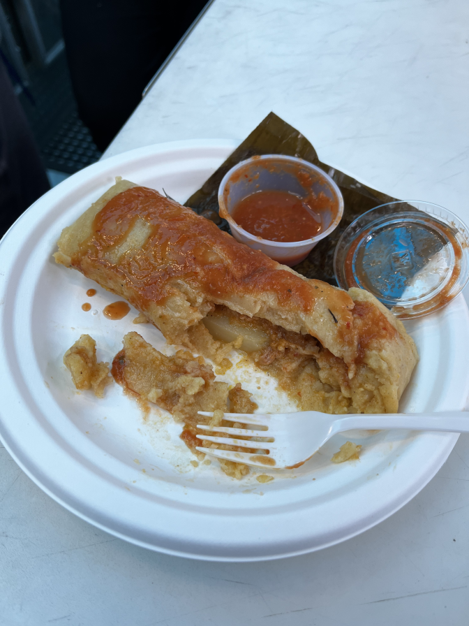 A half eaten tamale sits on a white paper plate next to a plastic fork and a small container of salsa