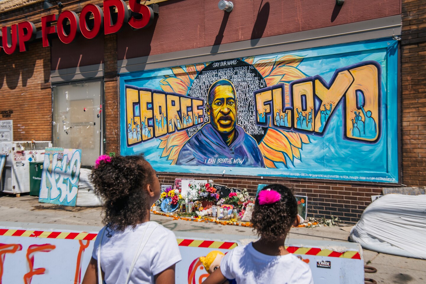 Two girls look at a mural of George Floyd, which displays his name and likeness against a blue background with a black and yellow sunflower.