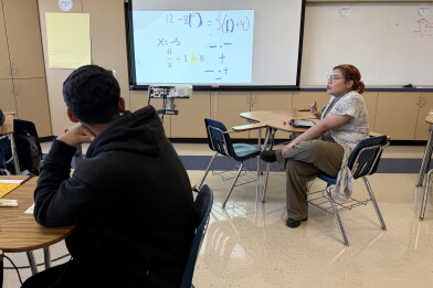 A teenage boy with medium skin tone and a black hoodie looks at a projection of math calculations. A women with medium light skin tone and glasses sits at a desk and gestures as though mid-speech. 