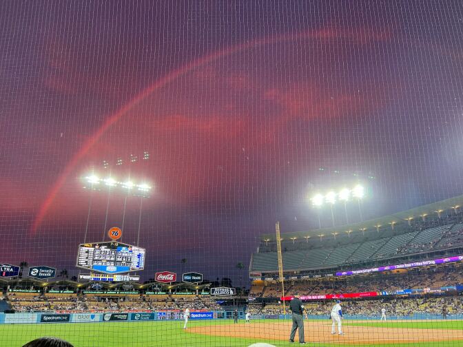 A salmon pink rainbow in a pink hued sky, seen from a baseball field. 