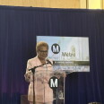 A woman with short brown hair stands in front of a podium with a microphone. The woman is wearing a light pink suit and a white shirt underneath. The podium is emblazoned with the letter "M" in white in a black circle. Behind the woman is a screen that has the same "M" logo and "Metro" written in black. There's a black banner at the bottom of the screen that reads"The Honorable Karen Bass" in all caps.