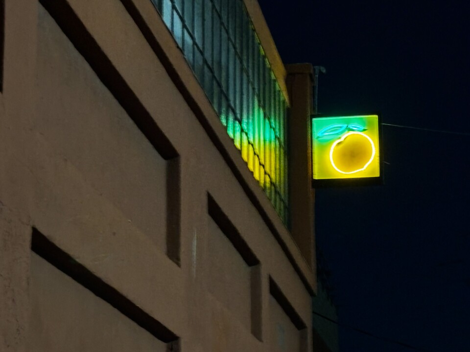  A glowing neon sign shaped like a yellow-orange fruit with green leaves is mounted high on the corner of an industrial building at night, casting colorful reflections on the glass windows beside it.