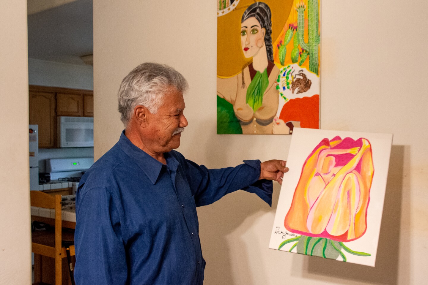 Arturo Gonzelez holds up one of his painting, a flower in reds, yellow and pinks. One the wall hangs another portrait of a woman with a long braid.