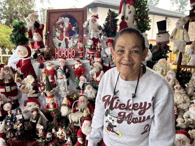 A woman with very short black hair is smiling while wearing a red and gray Mickey Mouse sweater and reading glasses on a chain around her neck. She's posing in front of a stand with dozens of Santas.
