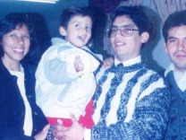 A slightly faded photo of a Latino man with glasses, smiling. holding a little boy with a jacket, red pants and brown hair. He is flanked by a smiling Latina with a bob haircut and a black jacket, and another man with dark hair and a faint mustache. 