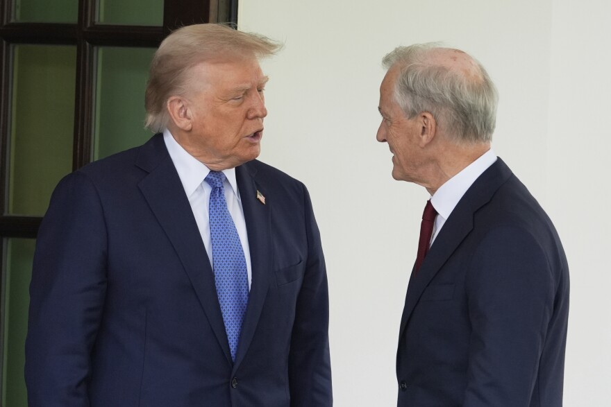 President Trump wearing a suit with a blue tie speaks to an older white man seen in profile.
