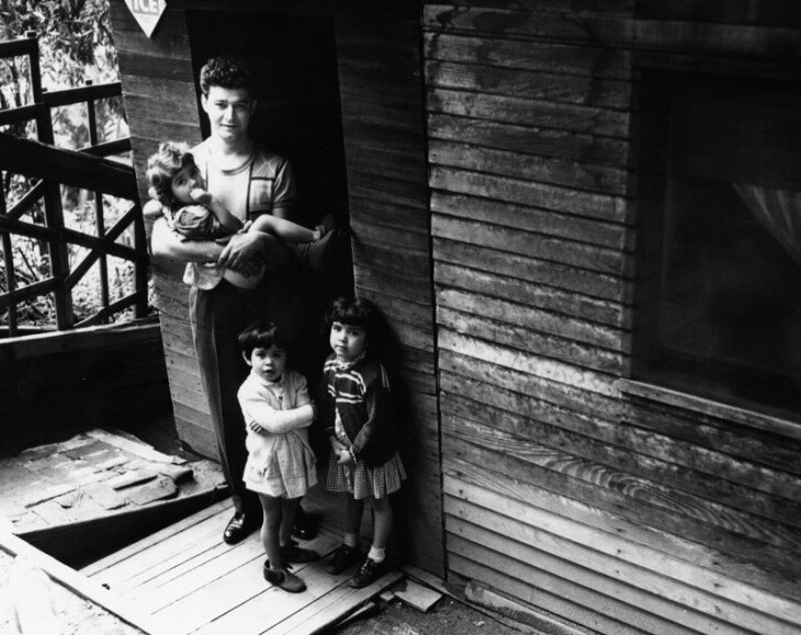 A man holds a toddler while two other small children stand nearby on a porch in a state of visible disrepair