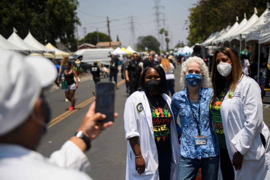 Barbara Ferrer wears her ID and a Dodgers mask, flanked by two health workers in white coats at a street fair in Watts, as another person takes a picture of them with a cell phone.