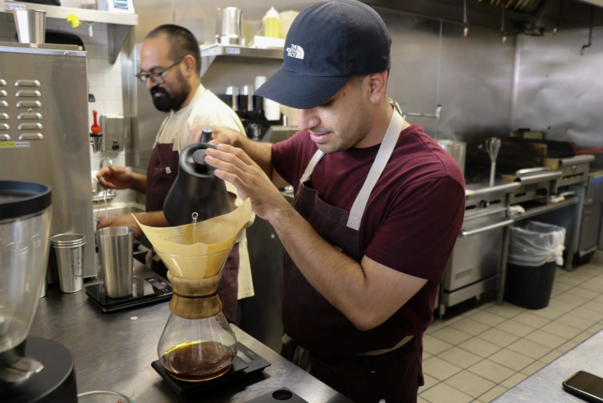 Two medium skinned men stand in a commercial kitchen. One is wearing a black baseball cap, a maroon T shirt and a black apron; he's pouring hot water through a coffee filter into a glass jug below. The other has glasses and a beard and is looking down as he rearranges something on the metal counter.