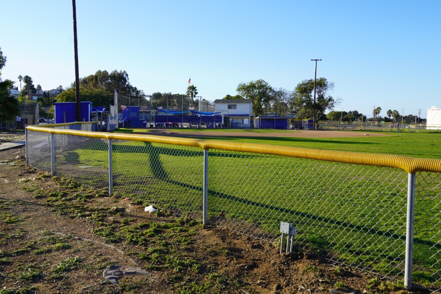 A wide view of the baseball field from behind the fence, which is yellow on top. In background you can see a building and blue bleachers.