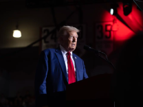 A man wearing a blue suit jacket and a red tie is standing behind a podium.